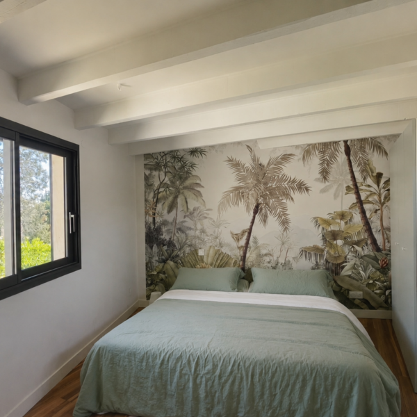 Chambre chaleureuse sous poutres peintes avec papier peint panoramique motif palmiers et jungle, lit double et fenêtre coulissante à Valras-Plage par Sophie Pico