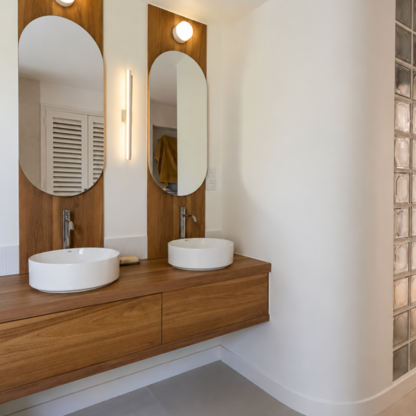 Meuble double vasque suspendu en bois sur-mesure avec miroirs ovales et cloison arrondie dans une salle de bains rénovée à Valras-Plage par Sophie Pico